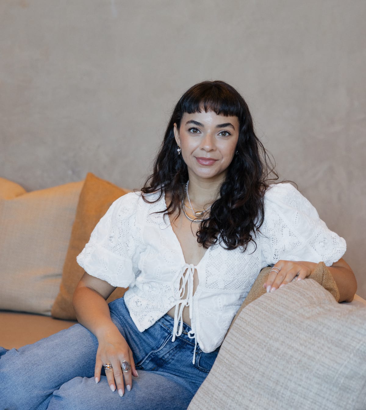 Eileen Silot seated on a sofa in a white lace blouse and jeans, one arm resting along the cushions, smiling gently at the camera.